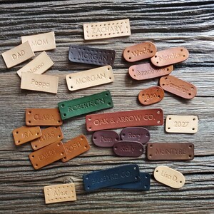May include: A collection of rectangular and oval leather tags in various colours including tan, brown, green, dark blue, and burgundy. Each tag is embossed with names or text such as ZACHARY, MOM, DAD, NANNA, POPPA, MORGAN, WADE, NATALIE, ANNABELLE, JENNIFER, BEN, CLARA, IRA, ELIAS, EMERY, JOSH, ROBERTSON, OAK & ARROW CO., PAM, RORY, KALLY, MCINTYRE, 2027, BLUEBYRD CO., and LISA D.