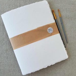 May include: A white, handmade paper journal with a brown paper band and a round sticker that says "Handmade by Paper Books".