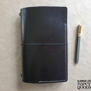 Puede incluir: Un diario de cuero negro con un cierre el&aacute;stico gris y costuras blancas. Un bol&iacute;grafo gris con punta dorada descansa junto al diario. El diario es rectangular y tiene esquinas redondeadas. El texto "GLORIOUS LIFE PAPER GOODS" est&aacute; en la esquina inferior derecha.