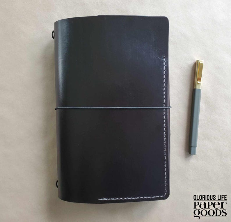 Puede incluir: Un diario de cuero negro con un cierre el&aacute;stico gris y costuras blancas. Un bol&iacute;grafo gris con punta dorada descansa junto al diario. El diario es rectangular y tiene esquinas redondeadas. El texto "GLORIOUS LIFE PAPER GOODS" est&aacute; en la esquina inferior derecha.