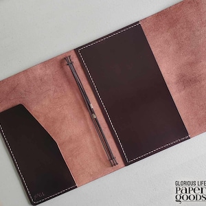 Puede incluir: Diario de cuero abierto con interior marr&oacute;n oscuro y exterior marr&oacute;n claro. El diario tiene un bolsillo en el lado izquierdo y un portal&aacute;pices. Las costuras blancas delinean los bordes. El texto "GLORIOUS LIFE PAPER GOODS" es visible.