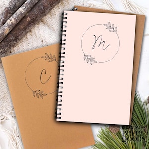 May include: Two spiral-bound notebooks with floral wreath designs. One notebook has the letter "M" inside the wreath, and the other has the letter "C". The notebooks are light pink and brown, with the text "GLORIOUS LIFE PAPER GOODS" at the bottom.