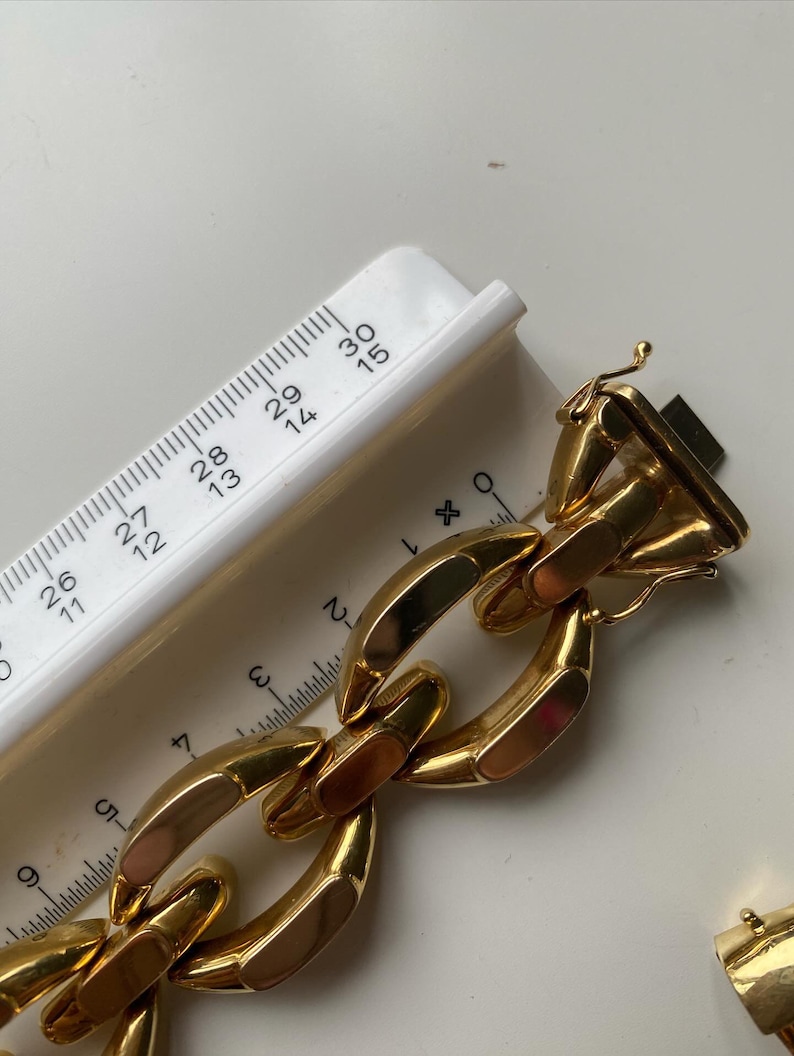 K&ouml;nnte beinhalten: Ein goldenes Kettenarmband mit gro&szlig;en, flachen, rechteckigen Gliedern. Das Armband ist etwa 6,5 cm breit und hat einen Verschluss.