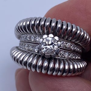 Può includere: Un set di anelli in argento con una grande gemma rotonda e trasparente al centro, circondata da gemme più piccole e trasparenti. L'anello ha un design unico e strutturato con un aspetto a coste. È un gioiello.