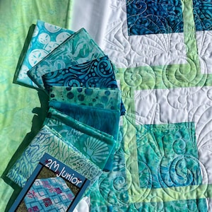May include: A quilt featuring a white and green grid pattern with blue and teal batik fabric squares. A stack of folded fabric swatches in shades of blue and green sits beside the quilt, accompanied by a 2M Junior pattern booklet with text.