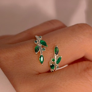 Puede incluir: Anillo plateado con diseño de hojas y gemas verdes esmeralda con corte marquesa. El anillo envuelve el dedo, con los elementos de hojas a ambos lados.