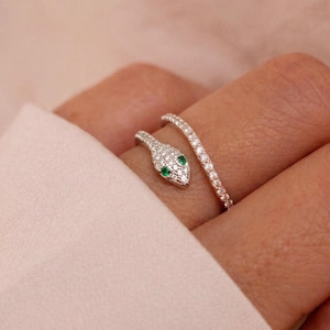 May include: A silver snake ring with a wrap-around design. The snake's head is encrusted with small, clear stones and features two green emerald eyes. The ring is worn on a finger, with the snake's head and tail wrapping around the finger.