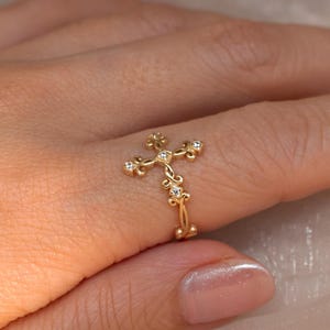 以下が含まれることがあります： 小さな透明な宝石がちりばめられた、十字架型のゴールドカラーのリング。リングは繊細で、上部と下部に花のモチーフが施された、細部までこだわったデザインです。指に着用されており、その精巧な職人技が際立っています。