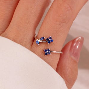 May include: A silver ring with a floral design. The ring features blue gemstones arranged in a flower and leaf pattern. The ring wraps around the finger, with the floral elements on top.