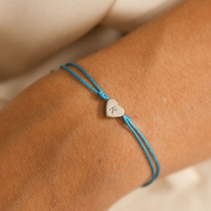 May include: A turquoise string bracelet with a silver heart charm engraved with the letter "K".