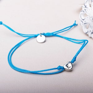 Peut inclure: Un bracelet en cordon bleu avec un pendentif en forme de cœur argenté et un pendentif en forme de disque argenté. Le pendentif en forme de cœur porte la lettre "E" gravée.