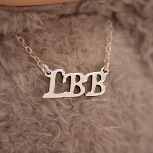May include: A silver-colored necklace with the letters "LBB" in a cursive font. The necklace is on a chain and rests against a soft, gray background. The letters are connected and have a polished finish.