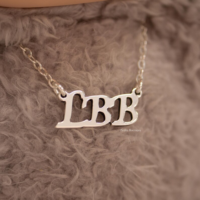 May include: A silver-colored necklace with the letters "LBB" in a cursive font. The necklace is on a chain and rests against a soft, gray background. The letters are connected and have a polished finish.