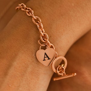 May include: A rose gold chain bracelet with a heart-shaped charm engraved with the letter "A".