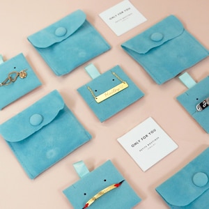 May include: Assortment of delicate gold necklaces and bracelets displayed on light blue velvet pouches. The jewelry features personalized names and a heart symbol. The pouches are arranged on a pink background with a small white card that reads "Only for you, Petite Boutique Jewelry".