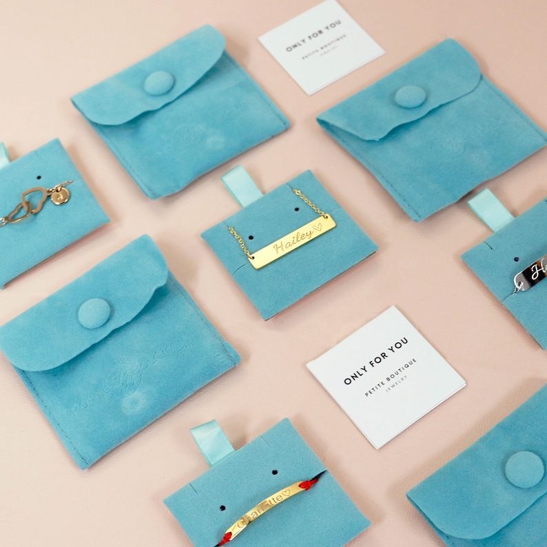 May include: Assortment of delicate gold necklaces and bracelets displayed on light blue velvet pouches. The jewelry features personalized names and a heart symbol. The pouches are arranged on a pink background with a small white card that reads "Only for you, Petite Boutique Jewelry".