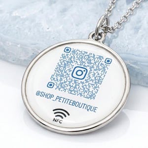 May include: A silver-colored pendant necklace with a white circular charm. The charm features a blue QR code with an Instagram logo, the text "@SHOP_PETITEBOUTIQUE", and the NFC symbol. The necklace chain is also silver.