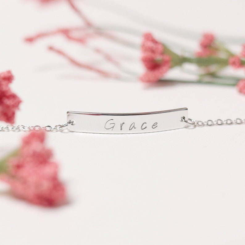 May include: Silver chain bracelet with a curved bar charm engraved with the name "Grace".
