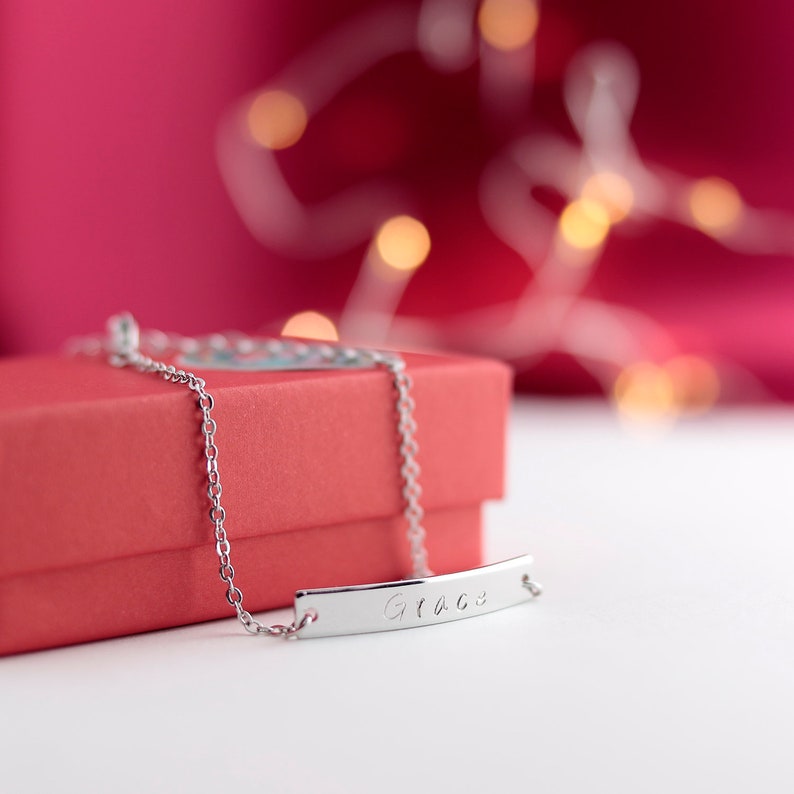 May include: A silver chain bracelet with a personalized bar charm engraved with the name "Grace". The bracelet is displayed on a red gift box.