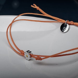 May include: A simple orange string bracelet with a silver daisy charm and a silver charm with the letter "S" on it.