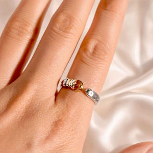 Peut inclure: Une bague ouverte argentée conçue pour ressembler à une ceinture miniature. L'anneau présente une boucle dorée à une extrémité et une série de petites indentations circulaires régulièrement espacées le long de la tige, imitant les trous de ceinture.
