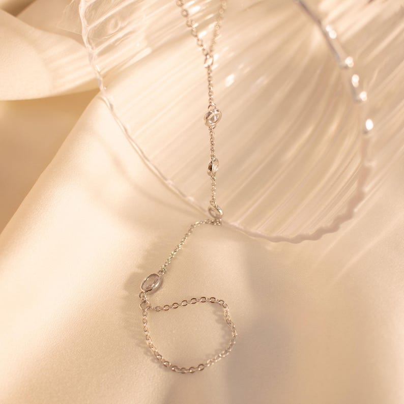 May include: A silver chain with small clear stones, connected to a ring. The chain is draped over a glass surface.