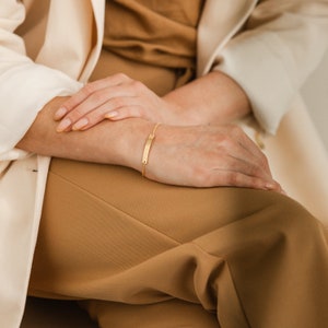 May include: A gold chain bracelet with a thin, rectangular bar charm. The bracelet is worn on a person's wrist.
