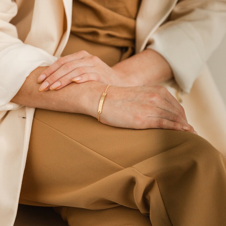 May include: A gold chain bracelet with a thin, rectangular bar charm. The bracelet is worn on a person's wrist.