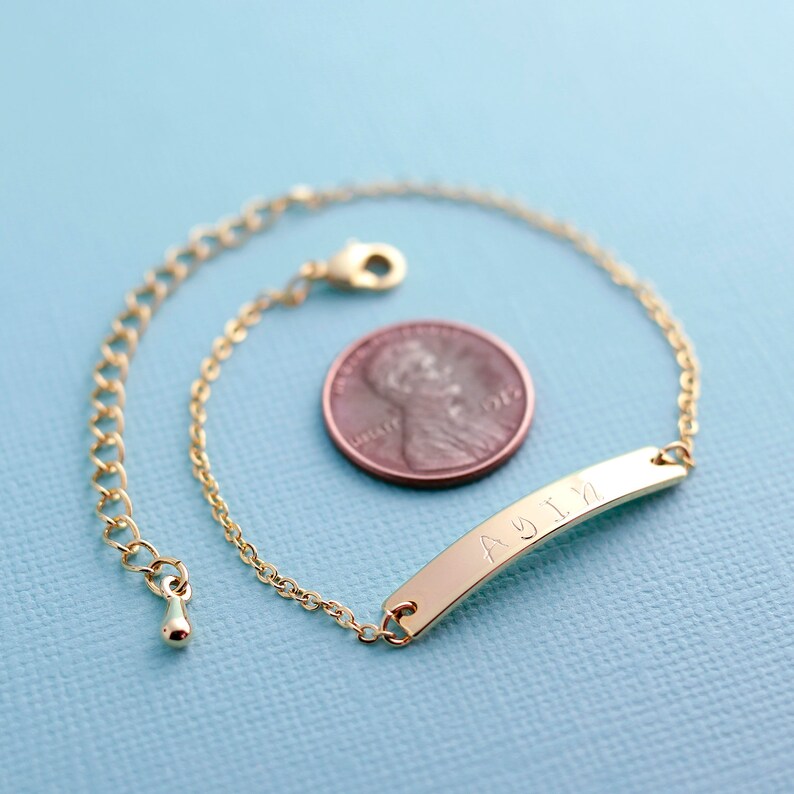 May include: A gold chain bracelet with a personalized bar charm engraved with the name "AYLA". The bracelet is shown next to a US penny for size reference.