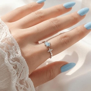 May include: A silver-toned open ring featuring a small, round, light blue stone set in a star-shaped frame on one end and a simple four-pointed star design on the other end. The band is thin and metallic, designed to be worn on a finger.
