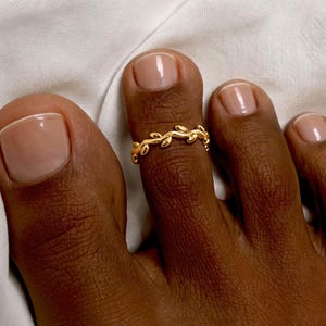 May include: A gold-toned toe ring featuring a continuous vine-like design with small, polished leaf-shaped elements. The ring is worn on the second toe, which has a light-coloured, neutral-toned manicure on the nail.