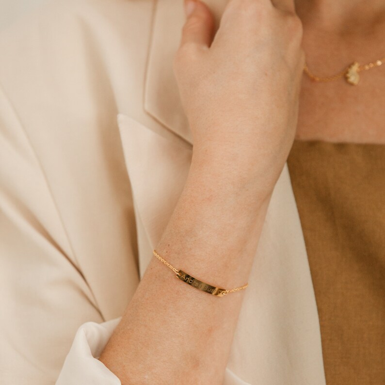 May include: A gold chain bracelet with a thin, rectangular tag that says "And So The Adventure Begins".