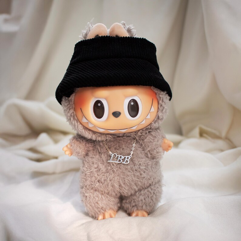 May include: A plush toy figure with a round, orange face, large black eyes, and a wide grin. It wears a black corduroy bucket hat and a furry gray outfit. A silver necklace with the letters "LBB" hangs around its neck.