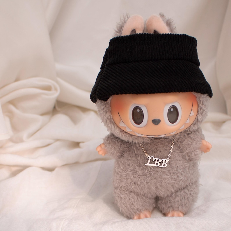 May include: A plush toy doll with a gray furry body, wearing a black corduroy hat and a silver necklace with the letters "LBB". The doll has large, round eyes and a smiling mouth.