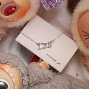May include: A silver necklace with the letters "LBB" is displayed on a white card that reads "Petite Boutique". The necklace is on a delicate chain. The background includes plush dolls.