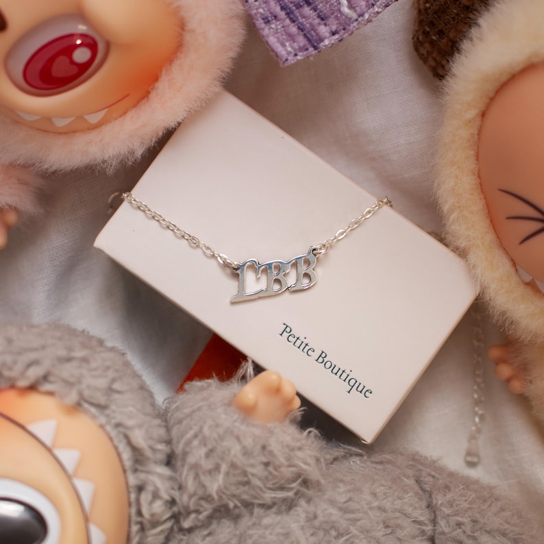 May include: A silver necklace with the letters "LBB" is displayed on a white card that reads "Petite Boutique". The necklace is on a delicate chain. The background includes plush dolls.
