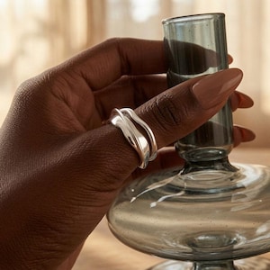 May include: A silver ring with a smooth, undulating, organic shape that wraps around the finger. The metal has a polished, reflective finish, creating soft highlights along its curved, flowing surface. The ring features a layered, double-band design.