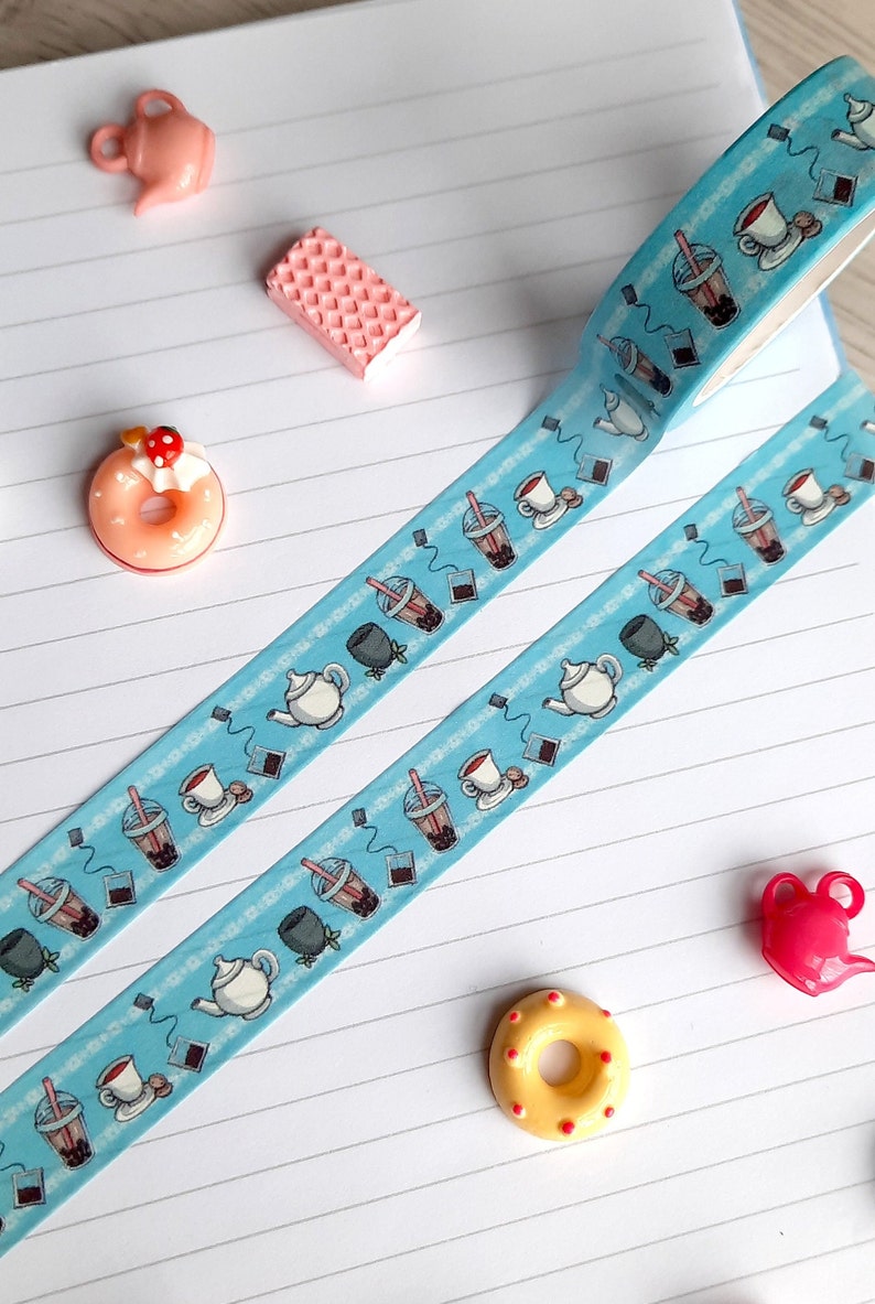 Pode incluir: Fita washi azul com um padr&atilde;o repetitivo de x&iacute;caras de ch&aacute;, bules e ch&aacute; de boba. A fita est&aacute; em um caderno branco com linhas e pequenos itens decorativos.