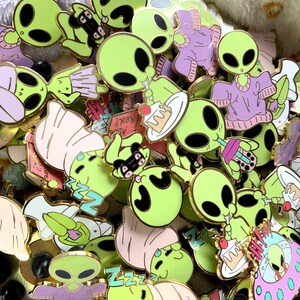 May include: A collection of colourful enamel pins featuring cartoon aliens in various poses. The pins are gold-toned and have a variety of designs, including aliens eating cake, drinking boba tea, and sleeping. Some pins have text, such as "Zzz" and "Alien's Realz".
