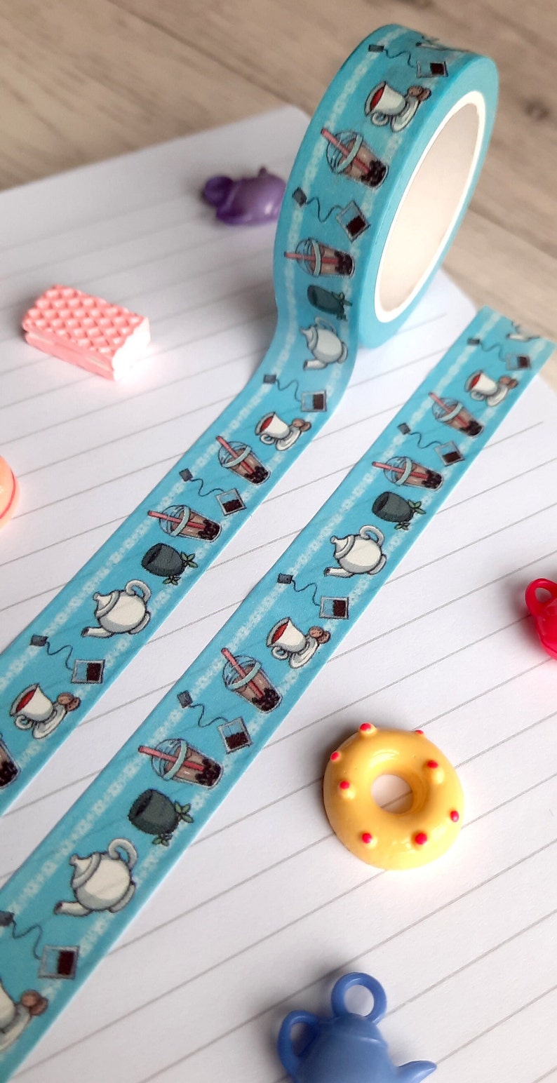 Pode incluir: Um rolo de fita washi azul com um padr&atilde;o repetido de x&iacute;caras de ch&aacute;, saquinhos de ch&aacute; e ch&aacute; de bolhas. A fita est&aacute; em um caderno branco com linhas.