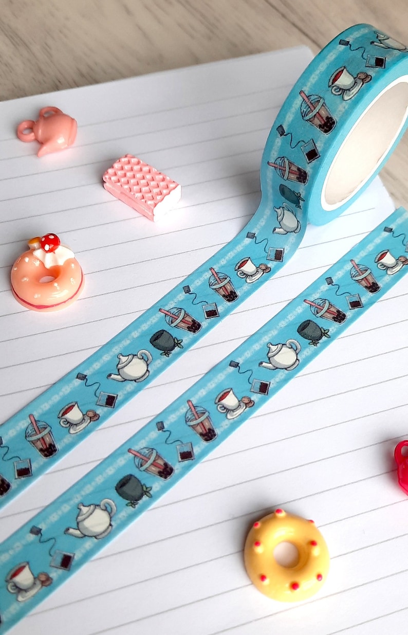 Pode incluir: Uma fita washi azul e branca com um padr&atilde;o repetido de x&iacute;caras de ch&aacute;, bules e ch&aacute; de boba. A fita est&aacute; em um caderno branco com linhas com donuts rosa e amarelos e uma bolacha de waffle rosa.