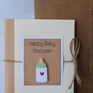 May include: A beige card with a brown paper insert. The insert has the text "Happy Baby Shower" and a white baby bottle with a green top and a red heart. The card is tied with twine.