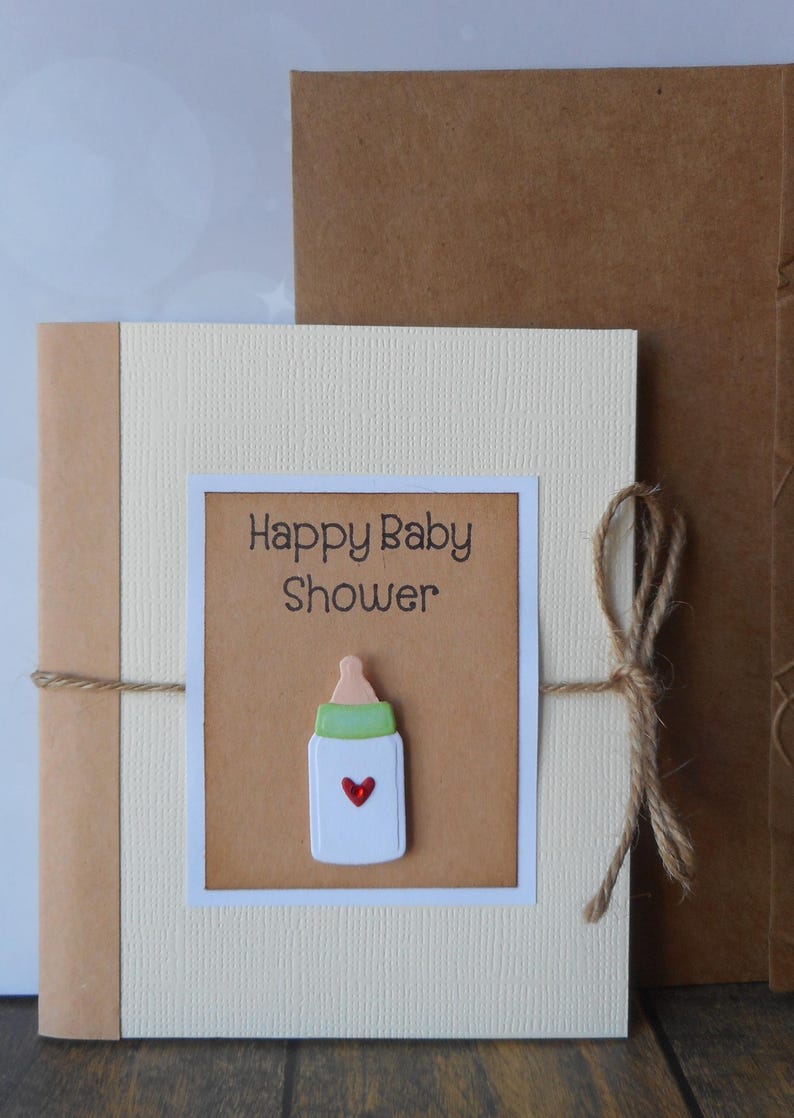 May include: A beige card with a brown paper insert. The insert has the text "Happy Baby Shower" and a white baby bottle with a green top and a red heart. The card is tied with twine.