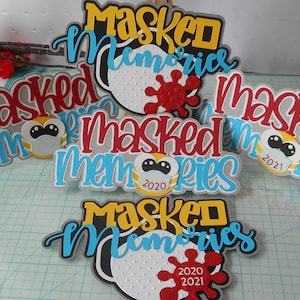 以下が含まれることがあります： 「Masked Memories」というフレーズをスタイリッシュなフェイスマスクと赤いウイルスグラフィックで描いた紙の切り抜きが5つあります。切り抜きは、赤、青、黄など、さまざまな色でできています。一部の切り抜きには、「2020」と「2021」のテキストが含まれています。