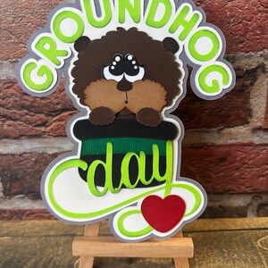 Peut inclure: Décoration de la fête de la marmotte avec une marmotte brune sortant d'un chapeau haut de forme noir avec une bande verte et noire. Les mots "GROUNDHOG DAY" sont en vert avec un contour blanc et un cœur rouge en bas.