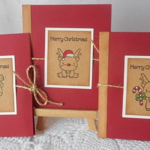 May include: Three red Christmas cards with brown paper inserts featuring illustrations of reindeer with different holiday accessories. Each card has a white border and the text "Merry Christmas!" printed on the insert.
