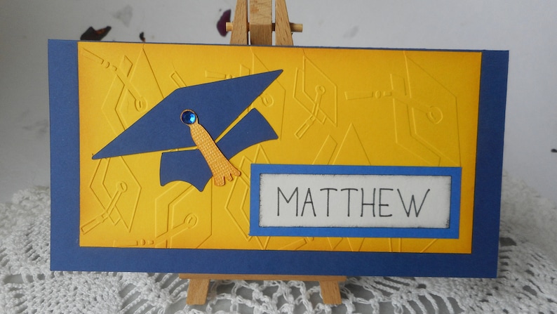 May include: A yellow card with a blue graduation cap and the name "MATTHEW" in a blue rectangle. The card has a blue border.