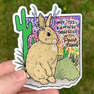 May include: A sticker depicting a tan jackalope with antlers, set against cacti and foliage. The text says "WHY YES, I AM ALSO OBSESSED WITH Pedro Pascal" on a glittery backdrop. The sticker has a white border.