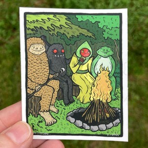 May include: A colorful illustration depicts a gathering of whimsical creatures around a campfire. A Bigfoot-like figure, a dark monster, a figure in a yellow robe, and a frog-like character are seated near the flames. The artwork is set in a forest scene.