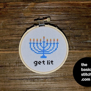 May include: A white embroidery hoop with a blue menorah with nine yellow and orange flames. The text "get lit" is embroidered below the menorah.